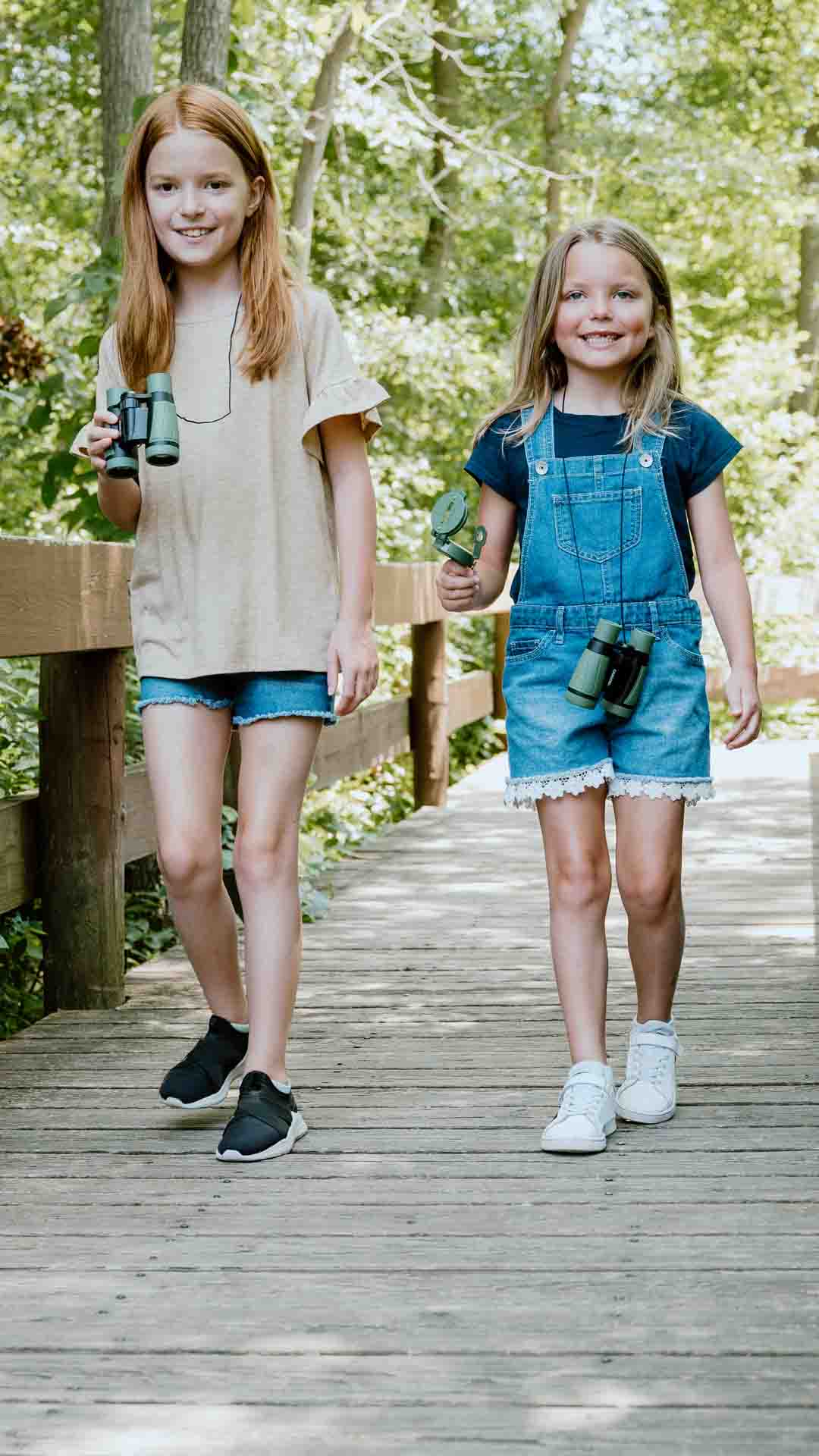 Two happy girls walking along a forest path each with a pair of binoculars